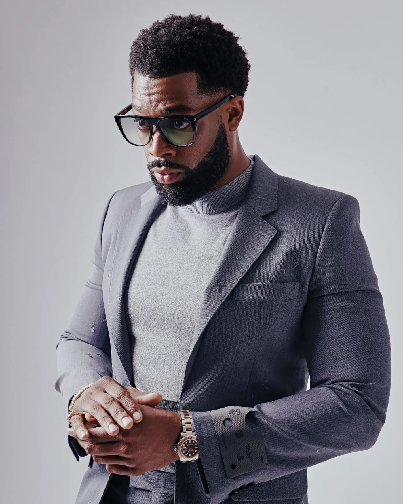 LaRoyce Hawkins in a grey suit and glasses, hands clasped, against a grey studio backdrop